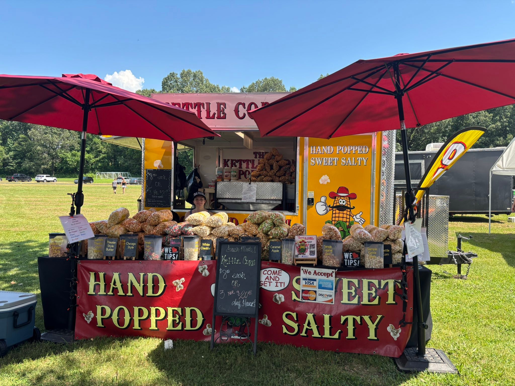 The Kettle Corn Poppers stand set up under red umbrellas with bags of kettle corn on display.